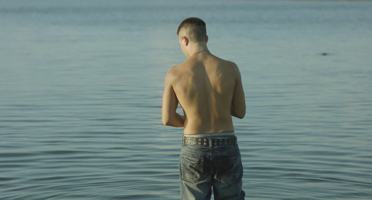 It Felt Like Love (2013) movie still: medium — A person stands waist-deep in calm water under bright natural daylight conditions. The shirtless man…