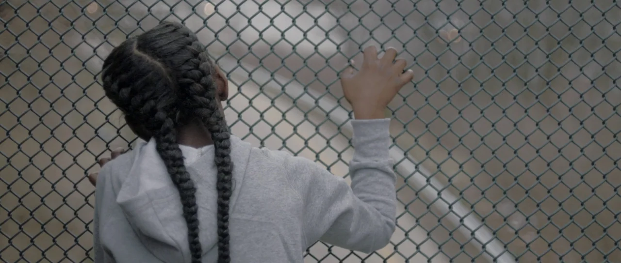 The Fits (2016) movie still: medium — A close view of a child standing behind a standard grey metal chain link fence. The girl holds the wires with…