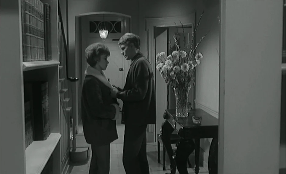 The Servant (1963) movie still: medium — A residential home entryway features a wooden side table and built-in bookshelves. A man stands facing a…