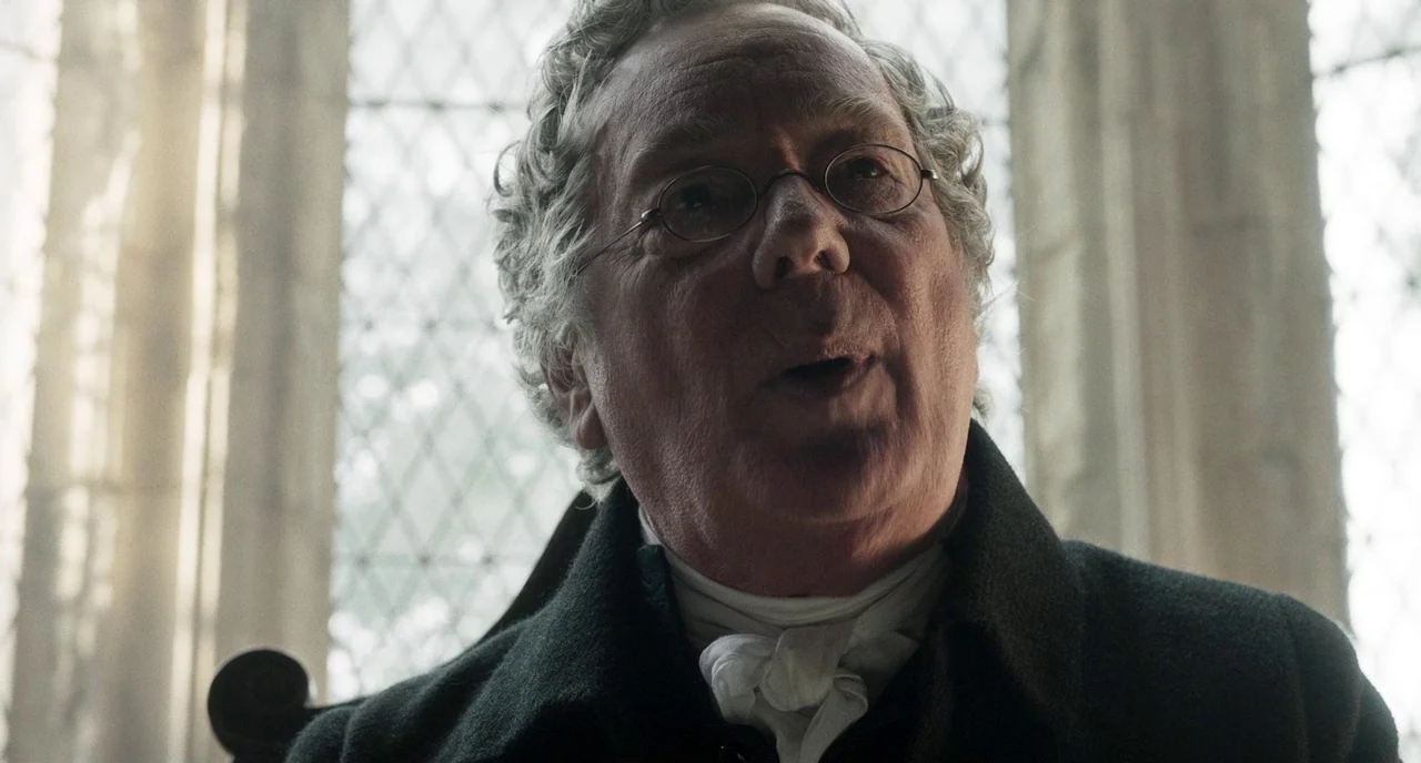 Peterloo (2018) movie still: closeup — A stone interior with large arched windows featuring diamond-pattern leaded glass panes. A man in period…