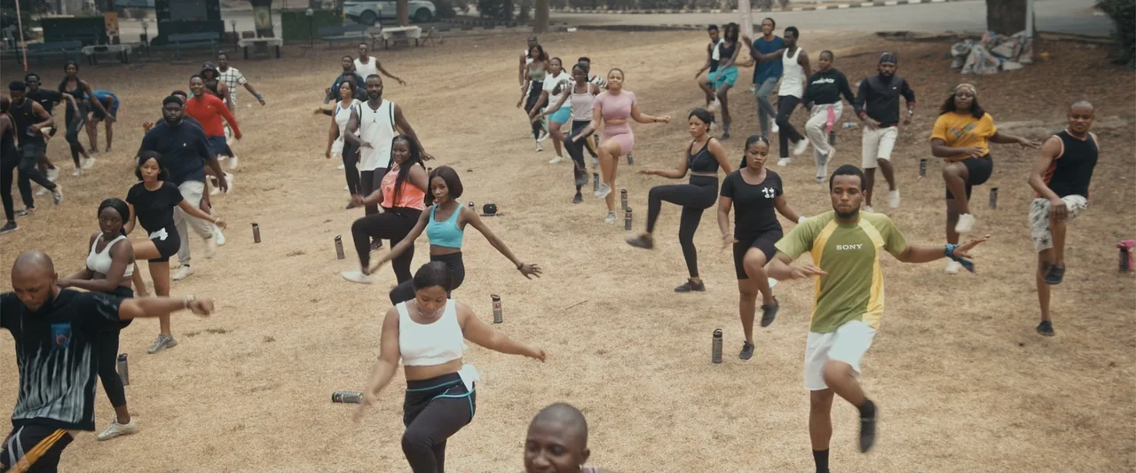 Big Love (2012) movie still: wide — A dry, grassy field serves as an outdoor fitness area for a large group. Many men and women in athletic apparel…