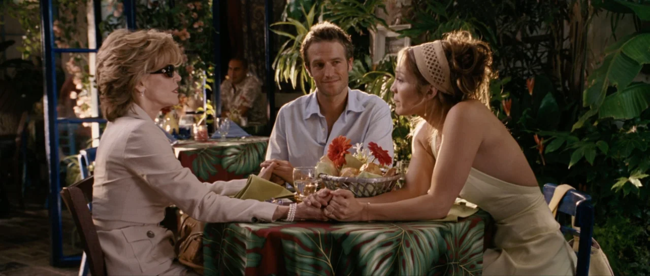 Monster-in-Law (2005) movie still: medium — A table set with floral patterned tablecloth is surrounded by dense indoor plants. Three people sit…