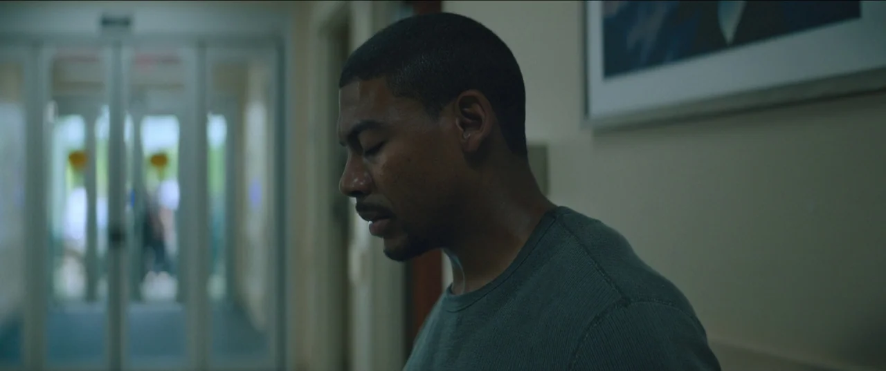 Rebel Ridge (2024) movie still: medium — A hospital corridor features white walls and a blurred glass doorway in the background. A man stands in the…