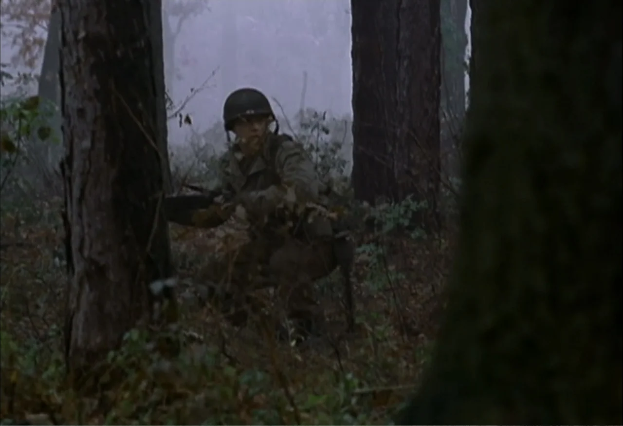 When Trumpets Fade (1998) movie still: medium — A dense, misty woodland setting filled with large pine trees and uneven forest terrain. A single…