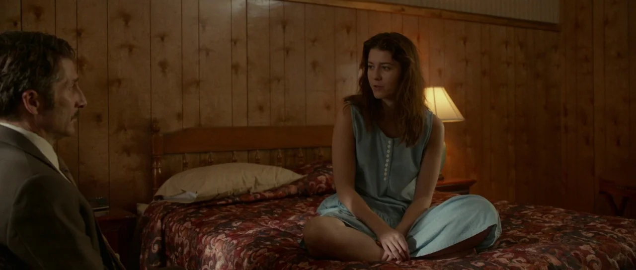 Faults (2014) movie still: medium — A motel room features vertical wood paneling and a bed with a floral bedspread. A man in a suit faces a young…