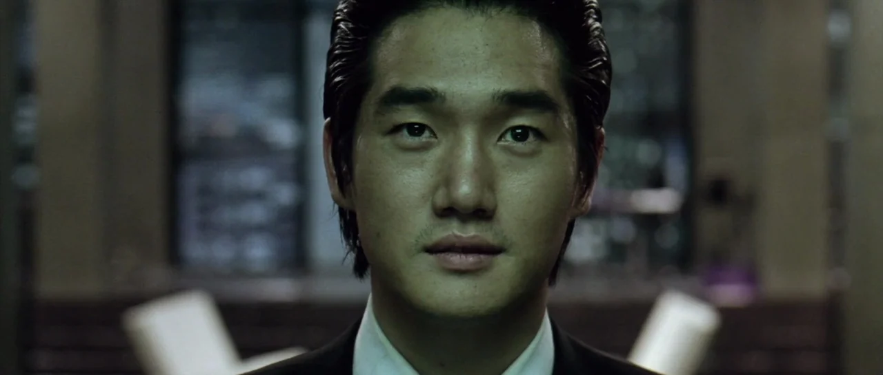 Oldboy (2003) movie still: closeup — A dark indoor space contains a man centered in the middle of the frame. The subject stands against a blurred…