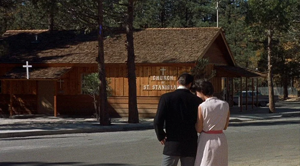 Kid Galahad (1962) movie still: medium — A wooden church building with a brown shingle roof sits beside a paved road. A man in a suit and a woman in…