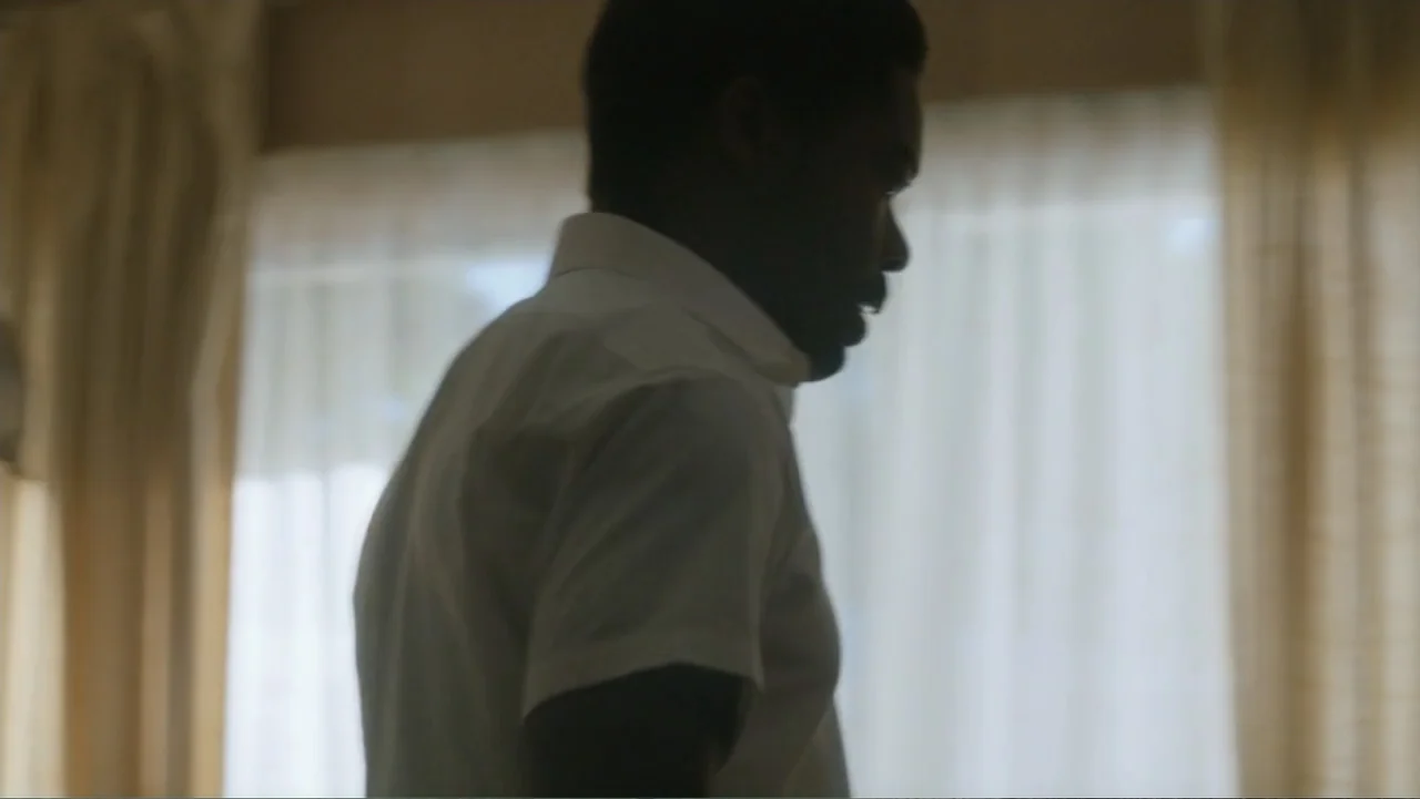 Nightingale (2014) movie still: medium — A man stands indoors near a bright window draped with translucent white curtains. The man wears a…