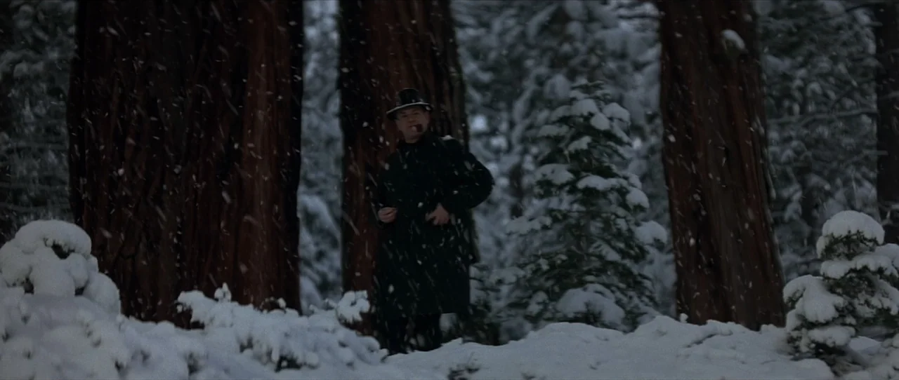 Cobb (1994) movie still: medium — A snowy woodland area filled with tall redwoods and fresh snow on the ground. A man in a dark coat stands between…