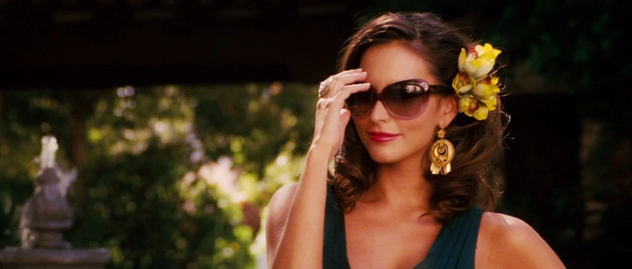 Casa De Mi Padre (2012) movie still: medium — An outdoor patio setting features a stone water fountain blurred in the background. A woman adjusts…