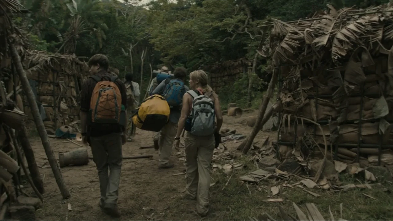 The Dinosaur Project (2012) movie still: fullbody — A group of hikers navigates a path through a dense jungle camp. They walk past makeshift…