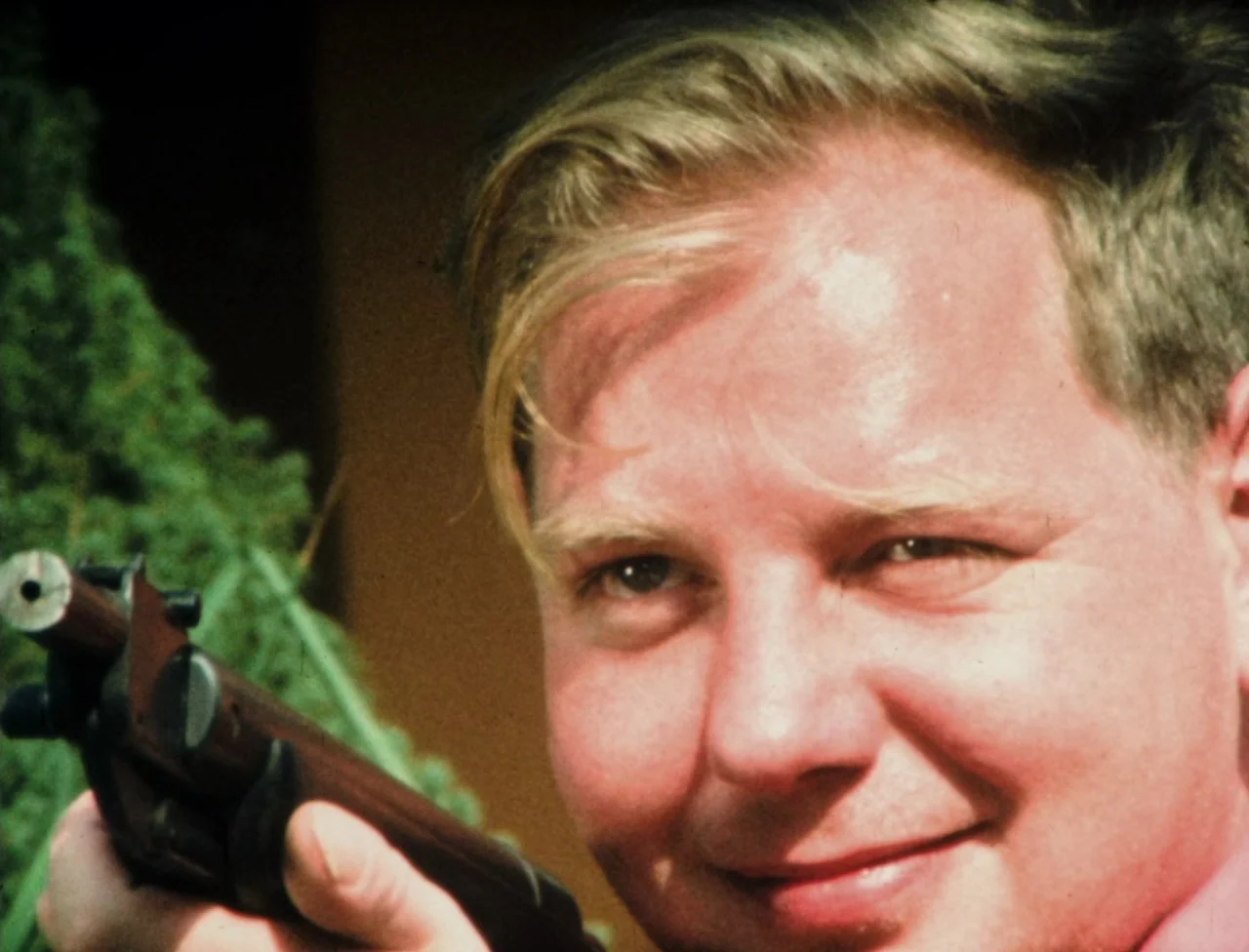 Nekromantik (1988) movie still: closeup — A man holds a rifle while positioned outdoors in front of a green coniferous shrub. The frame focuses on…