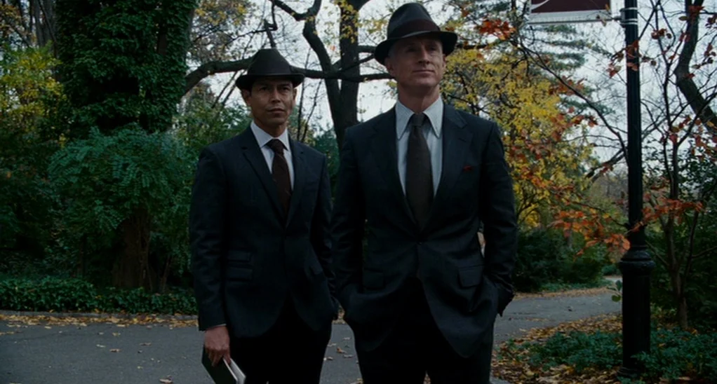 The Adjustment Bureau (2013) movie still: fullbody — Two men in business suits stand side-by-side on a paved park walkway. Autumnal trees with…