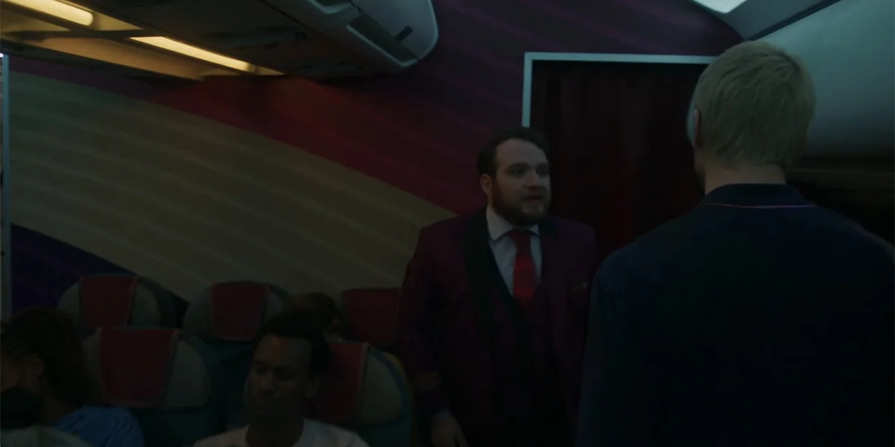 Fight or Flight (2025) movie still: medium — The interior cabin of a commercial airplane features rows of seats with purple headrests. A man in a…