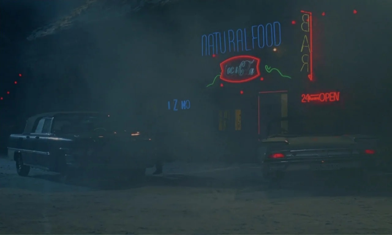 Ga-ga: Glory to the Heroes (1986) movie still: wide — A dark outdoor space contains two classic 1960s automobiles parked before a building wall.…