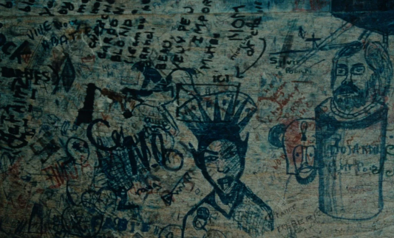 Graduate First (1978) movie still: closeup — A wooden wall is densely covered with amateur ink drawings and handwritten text. The surface shows a…