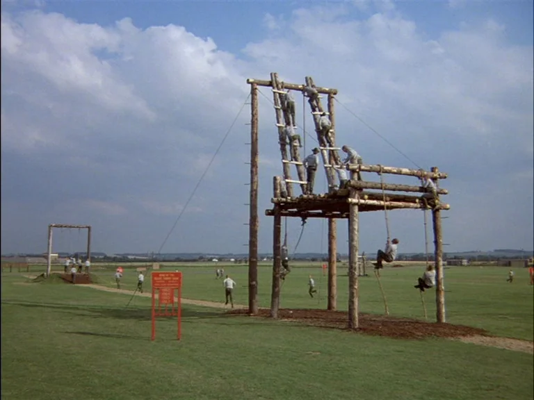 Full Metal Jacket (1987) movie still: wide — A vast grassy field contains a large, multi-level wooden obstacle climbing structure. Recruits in grey…