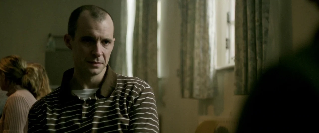 The Cured (2017) movie still: medium — A spare, neutral room with patterned curtains draped over a window. A man sits in the foreground, facing…