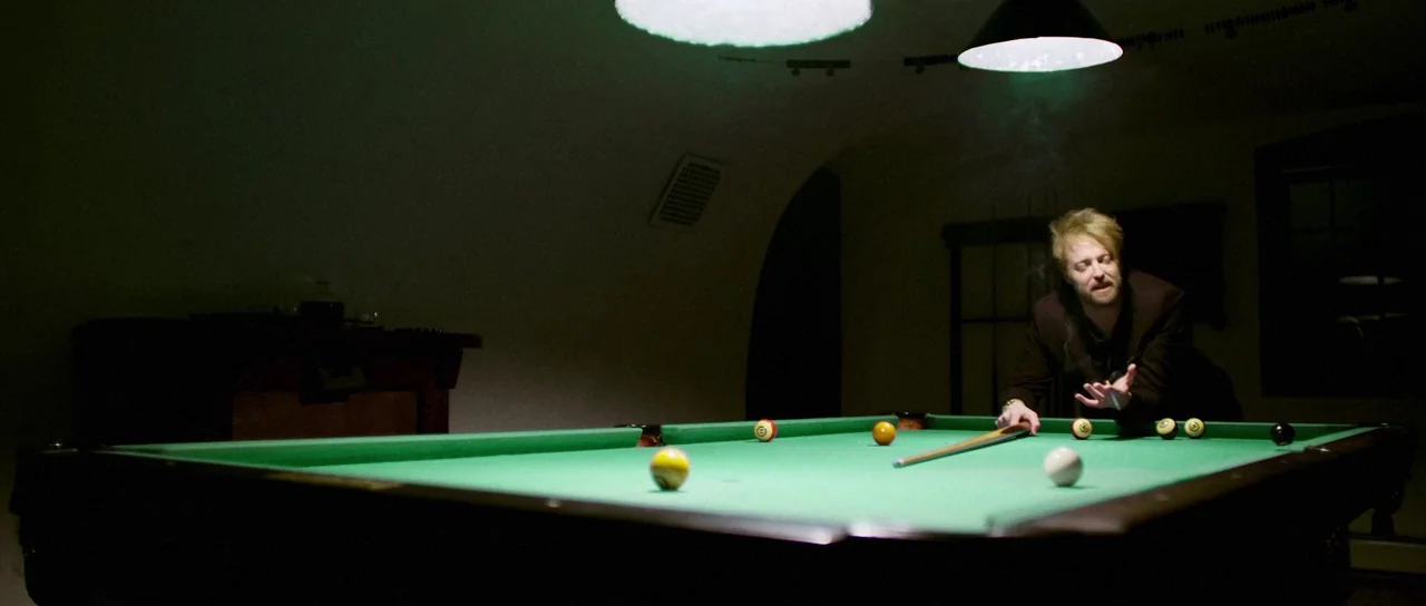 Depraved (2019) movie still: medium — A dimly lit room contains a green pool table centered in the frame. A man stands behind the table leaning over…