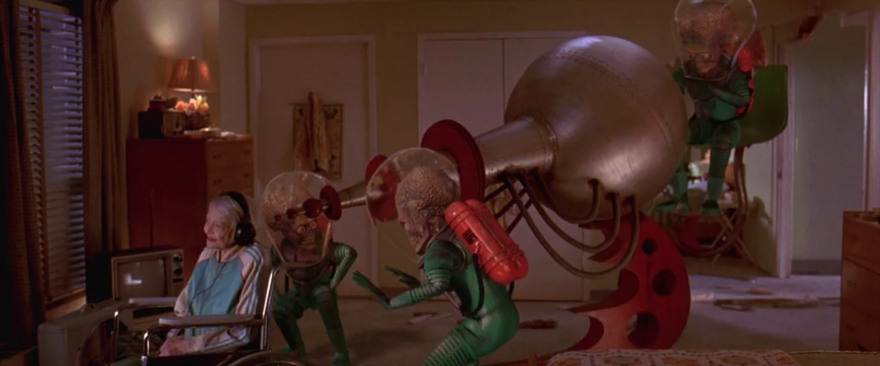 Mars Attacks! (1996) movie still: wide — A residential room contains an elderly woman sitting in a wheelchair wearing headphones. Three Martian…