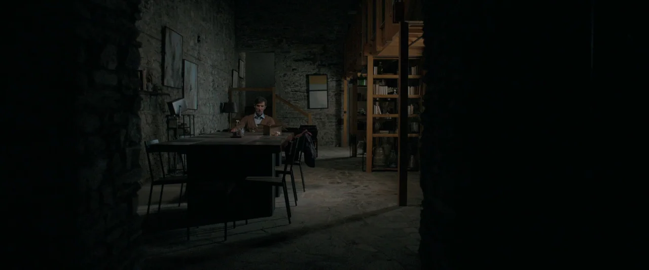Oddity (2024) movie still: wide — A spacious dining room with stone walls features a long table and wooden shelving. A man sits at the head of the…