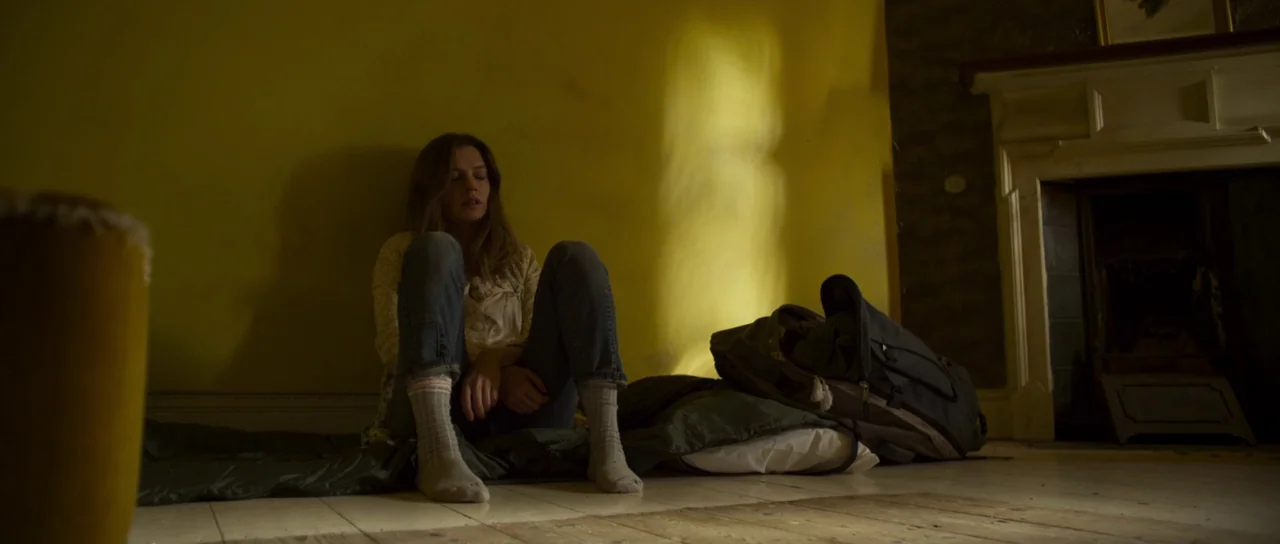 A Dark Song (2016) movie still: medium — A yellow-walled room with a white fireplace mantel and wooden flooring. A woman sits on a sleeping bag on…
