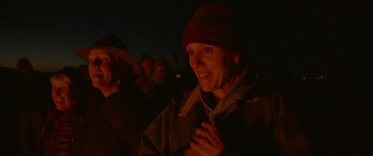 Nomadland (2020) movie still: medium — People stand together outside at night near an unseen fire source. The group members face slightly away from…