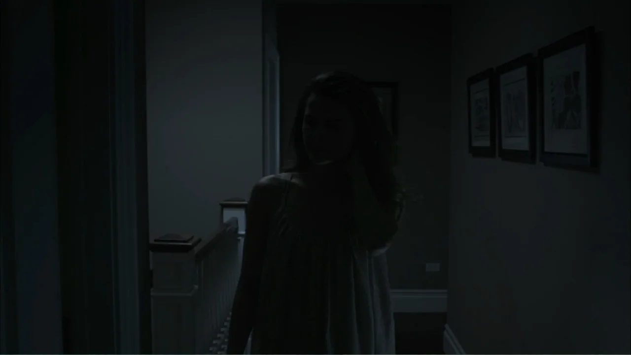 Dark Skies (2013) movie still: medium — A dark residential hallway features a white wooden banister and framed wall art. A woman stands in the…
