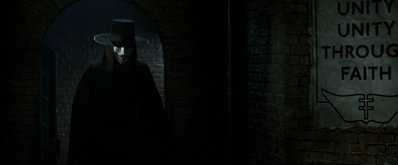 V for Vendetta (2006) movie still: medium — A dark brick tunnel features an archway with a single figure standing inside. A white poster with bold…