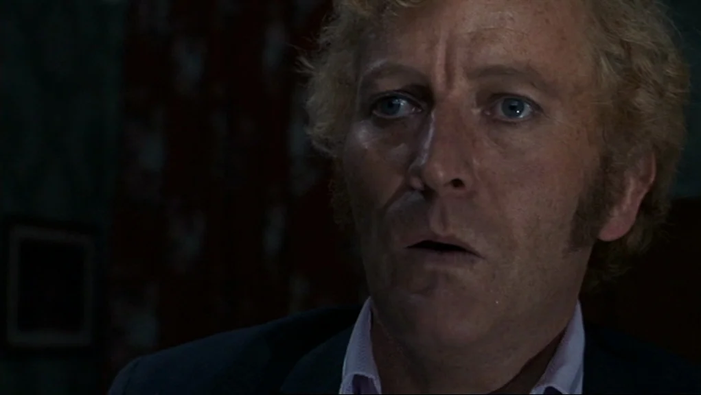 Frenzy (1972) movie still: closeup — A dimly lit room with dark wallpaper partially obscured by shadow. A middle-aged man stares intently with a…