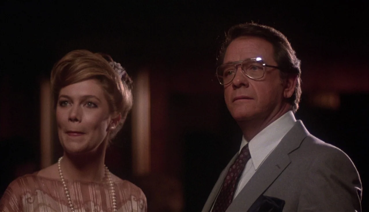 Body Heat (1981) movie still: medium — A dimly lit, formal interior space with dark walls and soft, directional light. A man and a woman in formal…