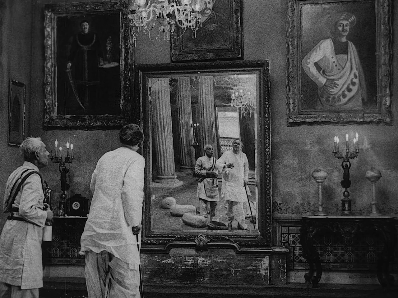The Music Room (1958) movie still: medium — A high-ceilinged mansion room features dark walls decorated with large oil paintings and candelabras.…