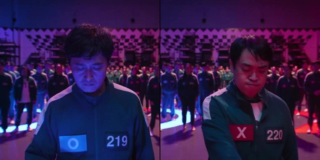 Squid Game (2024) movie still: medium — A split-screen interior view shows two men wearing green tracksuits in a large room. The background contains…