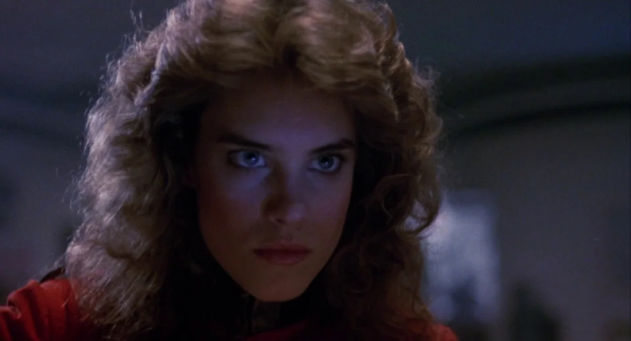 Night of the Comet movie still: closeup — A close-up shot of a young woman's face within a dimly lit room. She looks forward directly at the camera…
