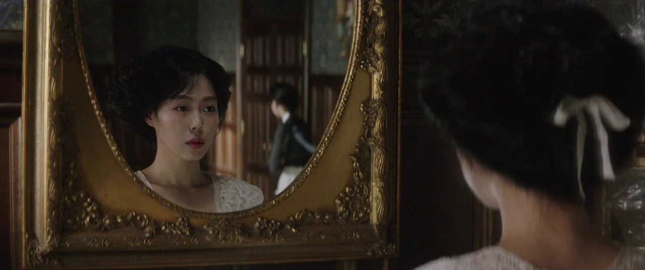 The Handmaiden (2016) movie still: medium — A woman stands before an ornate gold-framed oval mirror in a room with wood paneling. The reflection…