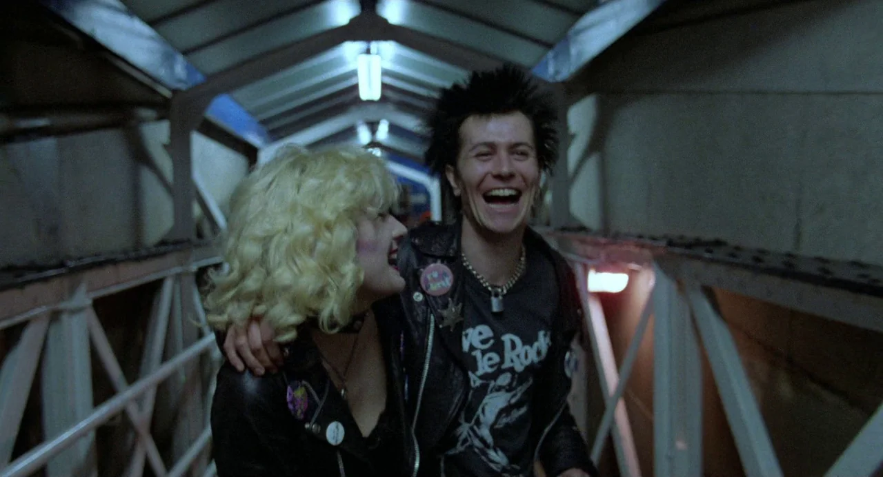 Sid and Nancy (1986) movie still: medium — A metal-framed pedestrian footbridge at night has glowing overhead fluorescent lights. A young man with…