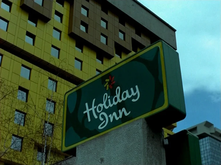 Notre Musique (2004) movie still: wide — A tall hotel building features a grid of yellow square windows and walls. A green rectangular Holiday Inn…