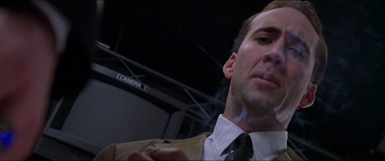 Snake Eyes (1998) movie still: closeup — A dimly lit room contains technical surveillance equipment and rack-mounted television monitors. A man in a…