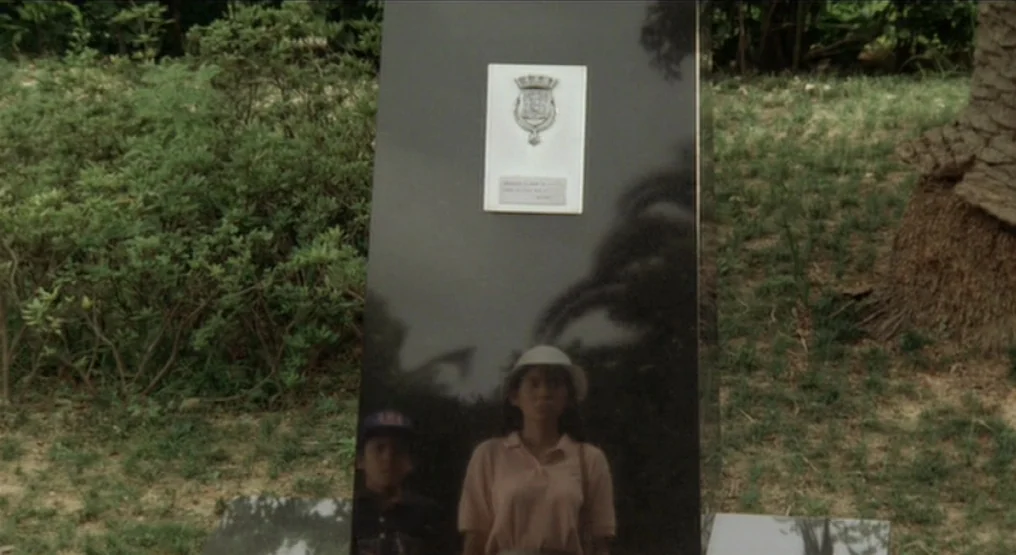 Rhapsody in August (1991) movie still: medium — A polished black stone monument stands in a grassy area with dense bushes nearby. A woman is seen as…