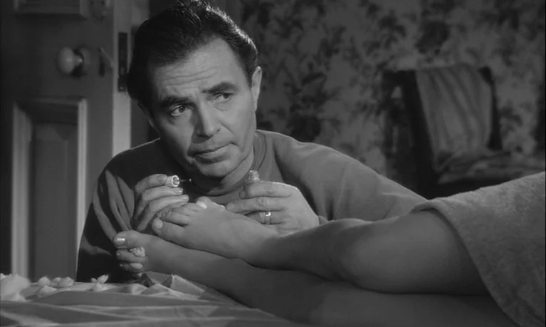 Lolita (1962) movie still: medium — A dimly lit room with floral patterned wallpaper serves as the backdrop. A man focuses on painting the toenails…