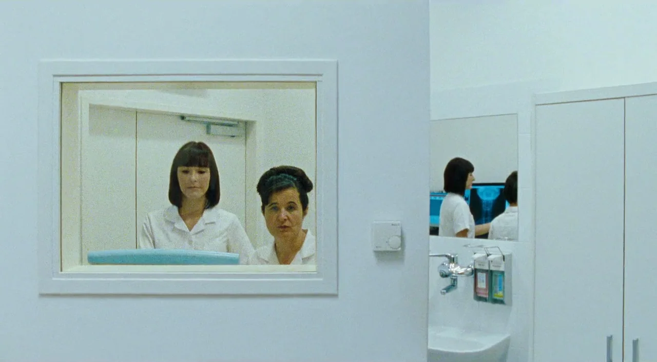 Paradise: Faith (2012) movie still: medium — A stark, minimalist white clinical room features a rectangular viewing window between two areas. Two…