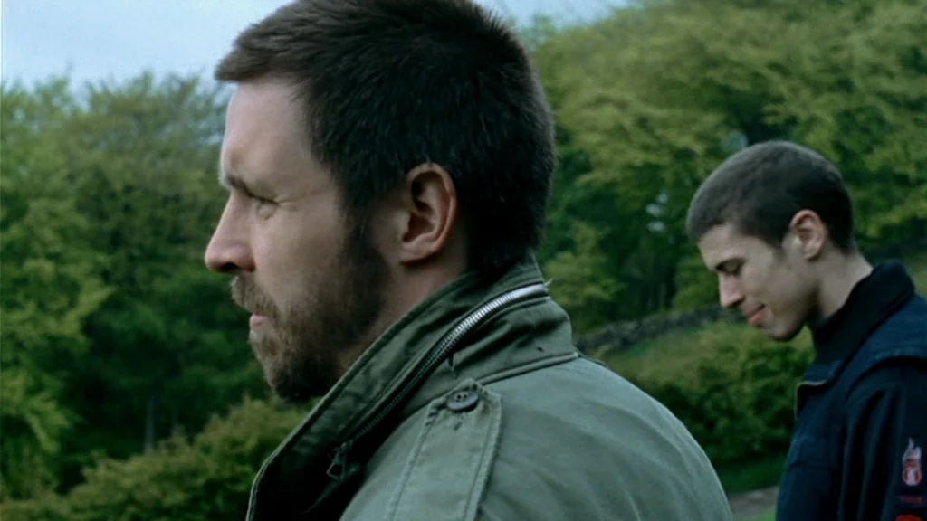 Dead Man’s Shoes (2004) movie still: medium — Two men stand in a dense green wooded area with mature trees. The foreground man looks left while the…