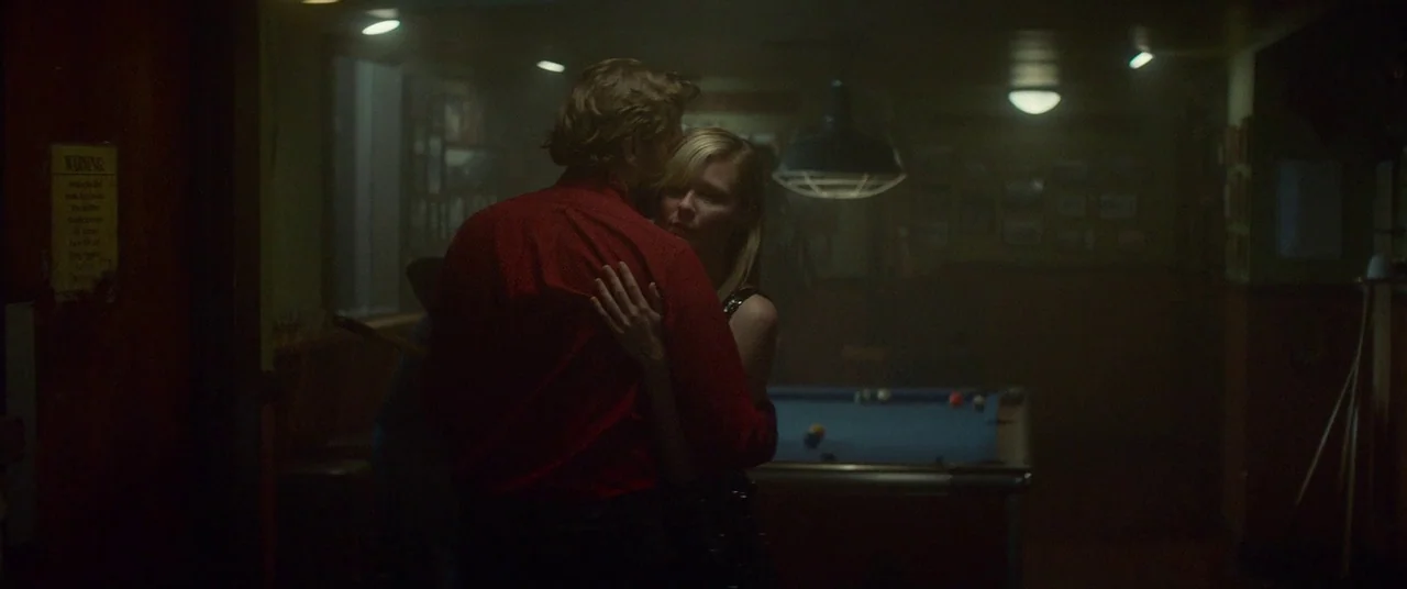 Woodshock (2017) movie still: medium — A dimly lit room contains a pool table and walls adorned with framed pictures. A man in a red shirt embraces…