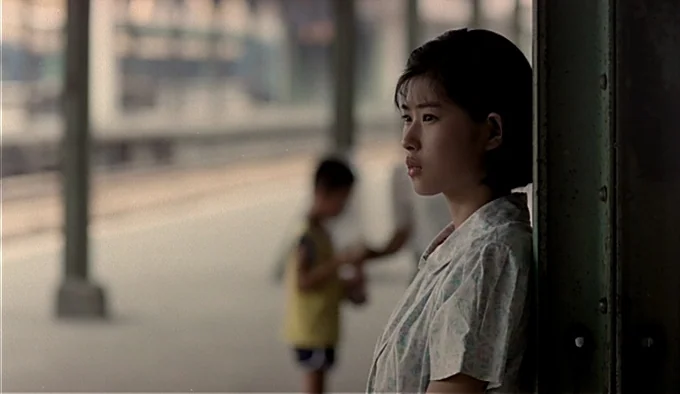 Dust In The Wind (1986) movie still: medium — An outdoor train station platform with a metal pillar in the foreground. A young woman leans against…