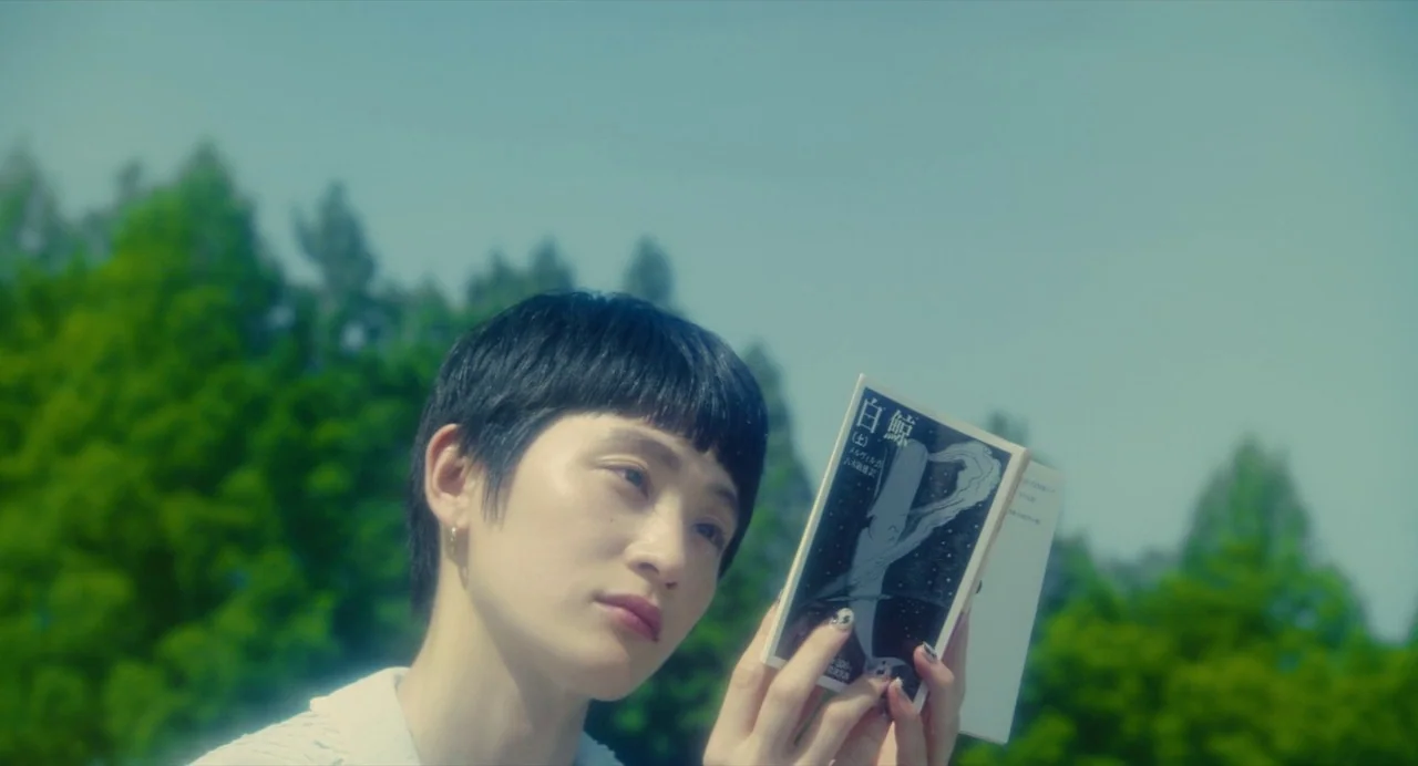 Diner (2019) movie still: closeup — A person stands outside in a park with blurred green trees in the background. She holds a small white book with…