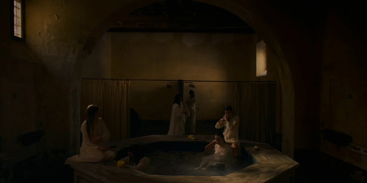 Immaculate (2024) movie still: wide — An octagonal stone pool sits in a dark, vaulted room with plaster walls. Several women in white garments bathe…