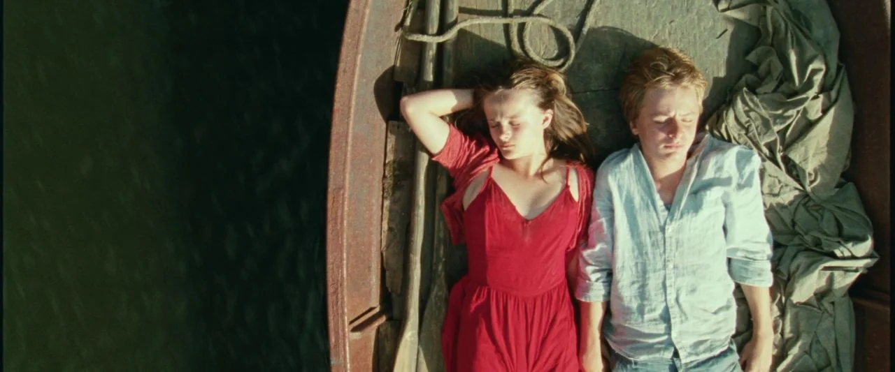 Adoration (2019) movie still: fullbody — A wooden rowboat floats on dark water in bright daylight. A young man and a young woman lie next to each…