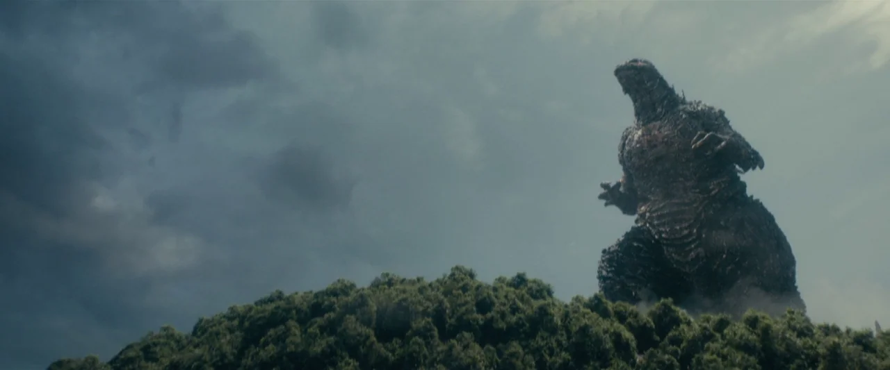 Godzilla Minus One (2023) movie still: wide — A colossal monster emerges from behind a dense green ridge under a stormy sky. The creature stands…