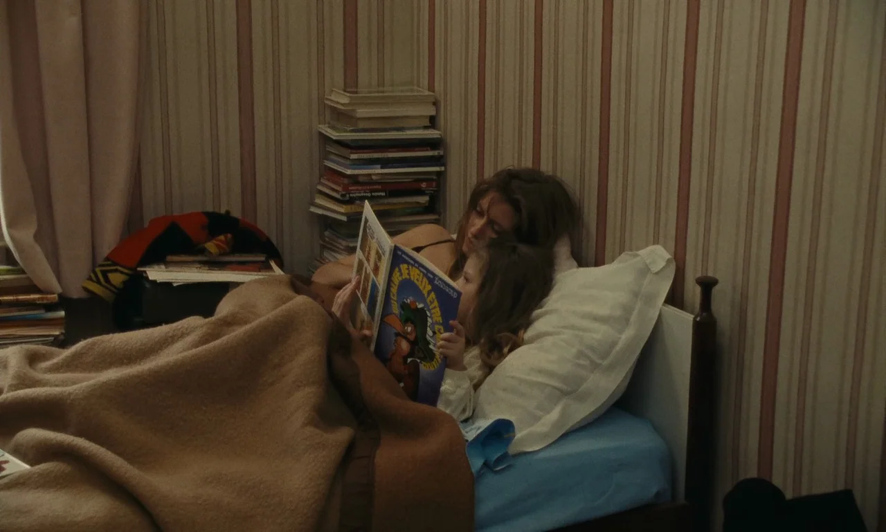 A Tale of Winter (1992) movie still: medium — A modest bedroom features striped wallpaper and a stack of books on a shelf. A woman and a small girl…