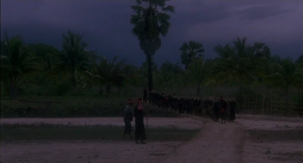 The Killing Fields (1984) movie still: wide — A line of people walks across a desolate, flat landscape at dusk. Palm trees stand as silhouettes…